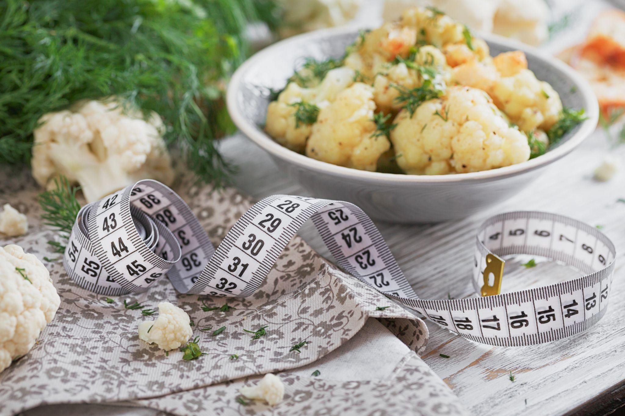 Low-Calorie Recipes A bowl of cooked cauliflower seasoned with herbs and oil, accompanied by a measuring tape symbolizing healthy eating for weight loss. Ideal for a clinic for weight loss or low-calorie meal inspiration