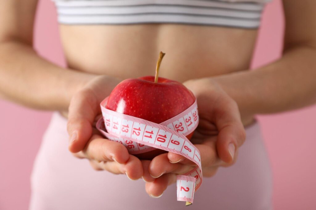 Close-up of a woman’s hands holding a red apple wrapped in a measuring tape, symbolizing mindful eating and healthy weight loss strategies. A great visual for weight loss clinic guidance