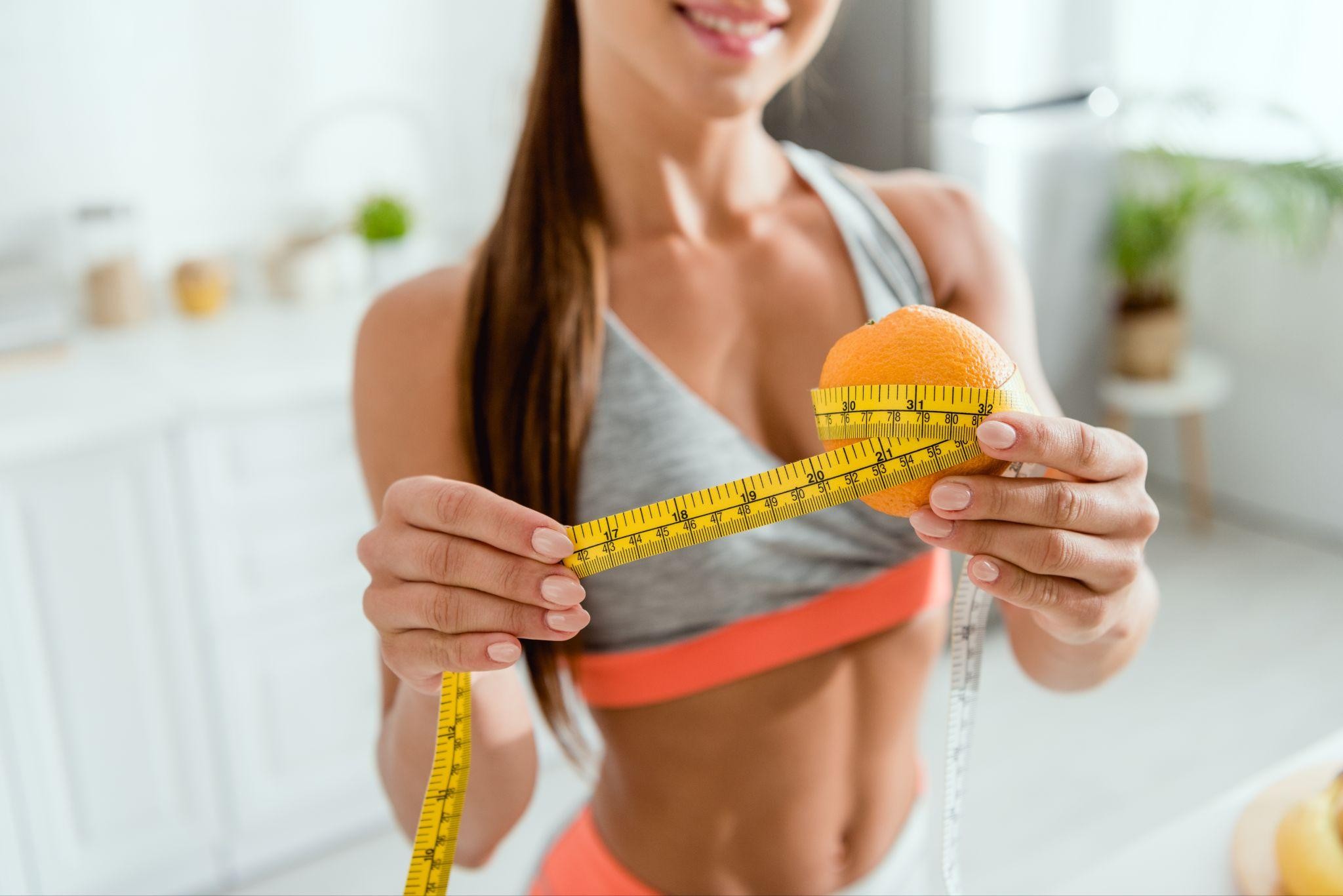 Rule for Weight Loss Fit woman in a sports bra smiling while measuring an orange with a tape measure, representing portion control and sustainable weight loss. Ideal for those searching for a clinic for weight loss and weight loss management near me.