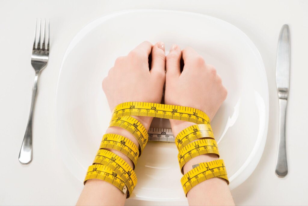 Hands tied with a yellow measuring tape over an empty plate with a fork and knife, symbolizing the struggle of dieting and extreme weight loss methods, related to reset metabolism fasting and Kirkland Weight Loss challenges.