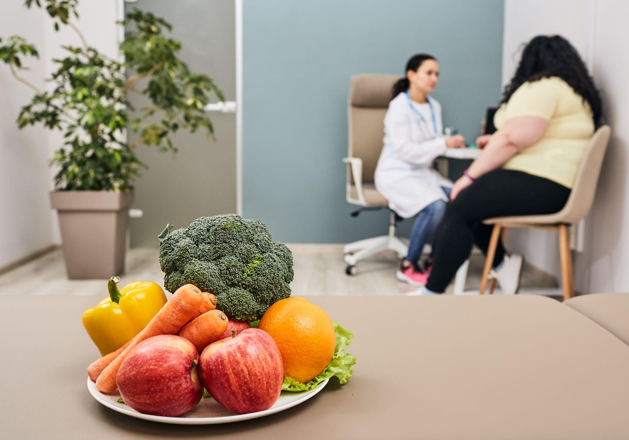 Eastside WL 1 Plate of fresh fruits and vegetables in a clinic setting, with a patient consulting a doctor in the background at a clinic for losing weight.